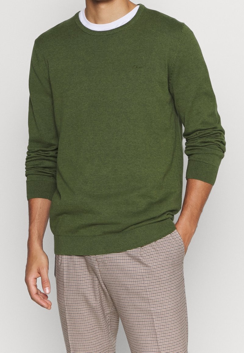 Homme portant un pull à manches longues vert olive sur un t-shirt blanc et un pantalon à carreaux marron clair, une main dans la poche.