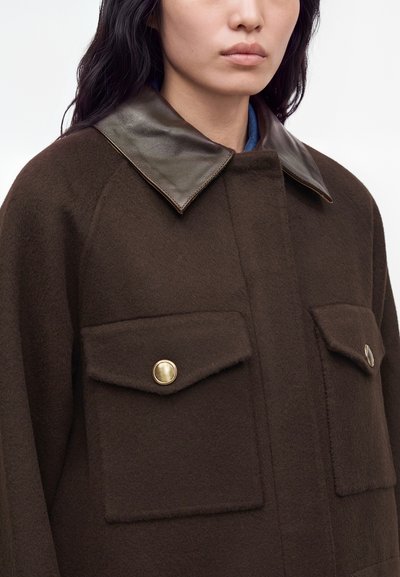 Femme portant un manteau en laine brun foncé avec de grandes poches avant et des boutons dorés, avec un col en cuir foncé brillant.
