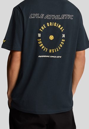Person trägt ein dunkles T-Shirt mit der Aufschrift "LYLE ATHLETIC" und kreisförmigem Text "THE ORIGINAL SCOTTISH LEAGUE 1874" sowie einer kleinen Fußballgrafik auf der Rückseite.
