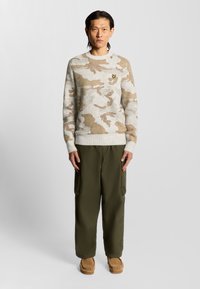 Hellgrauer Pullover mit einem beigen Camouflage-Muster und einem kleinen Logo, kombiniert mit olivgrünen Cargo-Hosen und hellbraunen Schuhen.