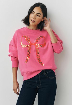 Lyserød sweatshirt med et stort, skinnende rødt bånddesign lavet af paletter. Rund halsudskæring og ribbede manchetter, parret med mørkeblå denimjeans.