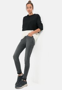 Zwart-witte geribbelde trui, slim-fit grijze jeans en zwarte sneakers met grijze accenten. Staand tegen een effen achtergrond.