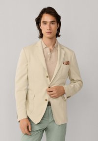 Mann trägt einen beigen Blazer mit gemustertem Einstecktuch, einem hellbraunen Poloshirt und hellgrünen Hosen, steht vor einem neutralen Hintergrund.