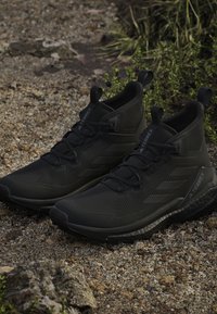 Ζευγάρι μαύρα πεζοπορικά παπούτσια Adidas Terrex σε βραχώδες έδαφος με πράσινα φυτά στο φόντο.