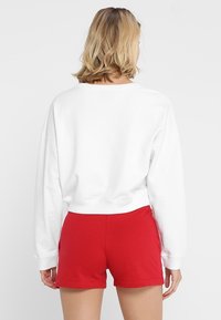 Sudadera blanca corta con escote ancho y mangas largas, combinada con pantalones cortos rojos. Material de algodón, diseño simple sin patrones.