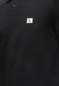 Μαύρο υφασμάτινο πόλο με ένα μαύρο κουμπί και μικρό λευκό λογότυπο με κόκκινα γράμματα «CK» και το κείμενο Calvin Klein στο στήθος.