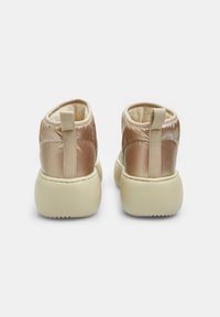 Botines acolchados en un brillante color oro rosa con forro de terciopelo beige. Presentan una tira en la parte trasera para ayudar a calzarlos y una suela gruesa beige con agarre texturizado.
