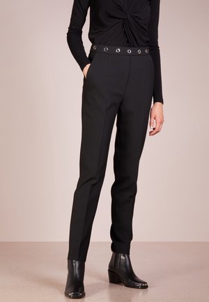 Kvinde iført tætsiddende sorte bukser med sølvfarvet øjlet bælte, sort langærmet vridd top og sorte ankelstøvler, stående med den ene hånd i lommen.