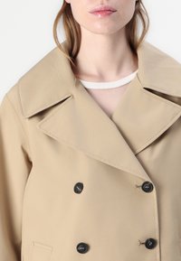 Beige dubbelknäppt kappa med skarp slagficka, svart knappdetalj, slät textur och subtil glans. Detaljer inkluderar en dold ficka.