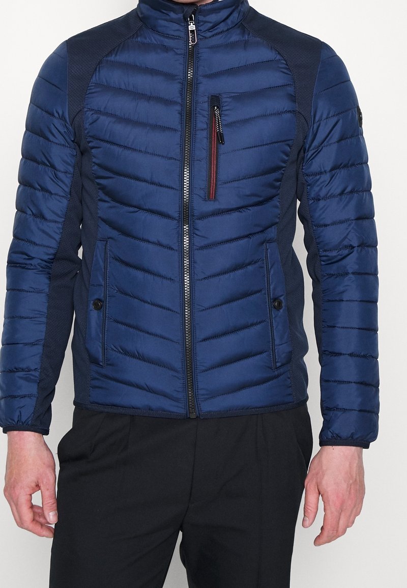 Ein Mann trägt eine taillierte blaue gesteppte Jacke mit Reißverschlusstasche auf der Brust und schwarze Hosen, steht vor einem weißen Hintergrund.