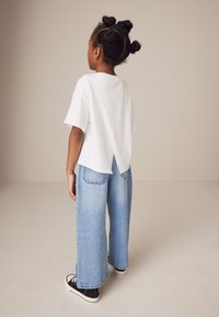 Hvid kortærmet skjorte med V-formet rygdetalje, parret med vide lyseblå denimjeans og sorte sneakers.