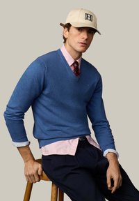 Mann mit beiger Kappe, blauem V-Ausschnitt-Pullover über rosa Hemd mit roter Krawatte, sitzt auf einem Holzschemel vor schlichtem Hintergrund.