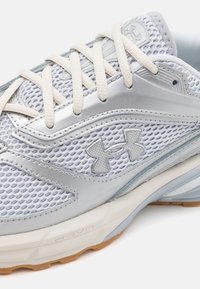 Gros plan d'une chaussure de course Under Armour blanche et argentée avec tissu en mesh, laçage frontal et semelle amortie marquée « HOVR ».