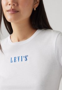 Hvit bomulls t-skjorte med blå brodert "LEVI'S"-logo, rund hals og korte ermer. Glatt tekstur med vanlig passform.