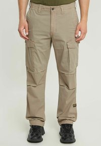 Pantaloni cargo beige con diversi taschini laterali, patta con zip, passanti per cintura frontali e una vestibilità ampia. Realizzati in tessuto leggero con una texture liscia.