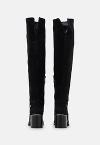 Botas altas de ante negro con caña partida, cremalleras en los laterales y un tacón ancho. Textura suave con detalles metálicos en la base del tacón.