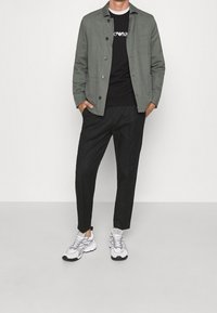 Giacca verde con bottoni, maglietta nera con grafica, pantaloni tapered neri e sneakers bianche con accenti grigi. Tessuti lisci.
