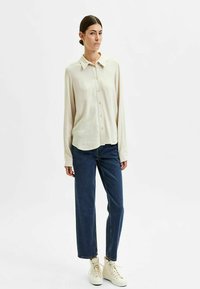 Licht beige blouse met kraag, voorkanten, en lange mouwen, gecombineerd met donkere blauwe rechte jeans en crèmekleurige sneakers.