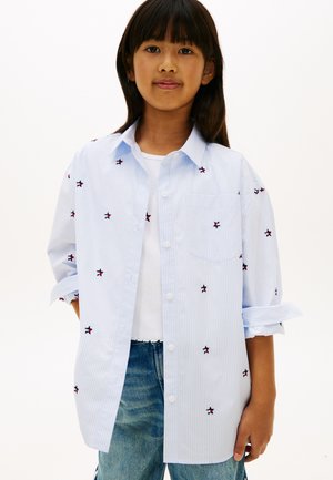 Fille portant une chemise à rayures bleu clair avec des boutons, ornée de petites étoiles brodées, par-dessus un top blanc et un jean bleu.