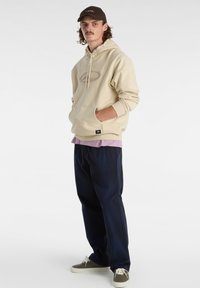 Felpa beige con cappuccio e tasca frontale, design ricamato, indossata sopra una maglietta viola chiaro. Pantaloni a scacchi scuri e sneakers verdi. Cappello marrone.