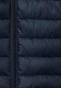 Giacca a vento blu navy con design trapuntato. Caratterizzata da una zip frontale e una superficie liscia e lucida. Leggera e isolante per garantire calore.