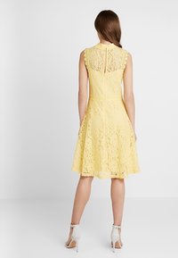 Žluté krajkové šaty s upnutým korzetem, rozšířenou sukní, výstřihem ve tvaru vlny a detaily ve tvaru klíčové dírky na zádech. Nošeny s bílými sandály na kotníku.