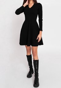 Robe longue à manches longues noire avec un col en V, un corsage ajusté et une jupe évasée. Associée à des bottes noires montantes lacées jusqu'au genou. Texture de tissu lisse.