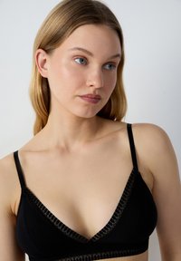 Brassière noire avec un décolleté en V profond, bordure en dentelle transparente, fines bretelles réglables et texture de tissu doux, présentant un design moderne et minimaliste.