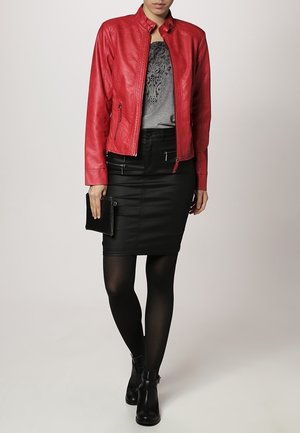 Femme portant une veste en cuir rouge, un haut en dentelle gris, une jupe noire, des collants noirs, des bottines noires, et tenant une pochette noire.