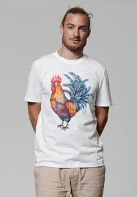 Weißes Baumwoll-T-Shirt mit einem bunten Grafikdesign eines Huhns, das detaillierte Federn und blaue Blattakzente aufweist, kurzer Ärmel und runder Hals.