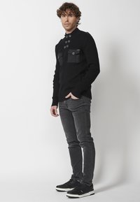 Czarny sweter prążkowany z metalowymi akcentami, dwie przednie kieszenie i wysoki kołnierz. W połączeniu z szarymi dżinsami i czarnymi sneakersami.