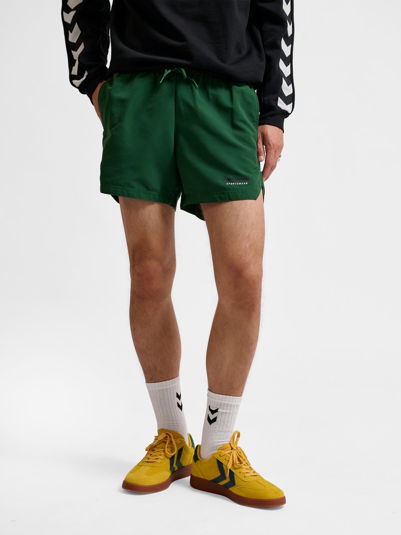 Mężczyzna ubrany w zielone krótkie spodenki sportowe, czarną bluzkę z długim rękawem z białymi paskami w kształcie szewronu, białe skarpetki i żółte trampki z brązowymi podeszwami.