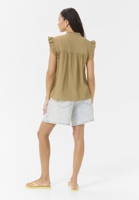 Blusa de textura verde oliva con mangas cortas y acampanadas, combinada con pantalones cortos de mezclilla azul claro y chanclas amarillas. Un atuendo simple y casual.