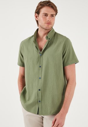Camicia a maniche corte, con bottoni, in tessuto verde chiaro con una finitura testurizzata. Presenta bottoni blu navy e un colletto classico, che arriva sotto la vita.