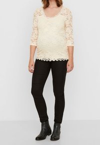 Lace top with three-quarter sleeves in light cream, featuring a scalloped hem and floral pattern. Paired with black fitted pants and ankle boots.