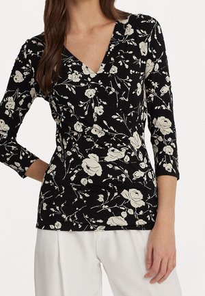 Kobieta ubrana w czarną, długorękawową bluzkę z dekoltem w kształcie litery V i białym kwiatowym nadrukiem oraz białe spodnie, stoi z jedną ręką zgiętą, a drugą zrelaksowaną.