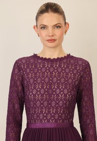 Violetti pitsimekko, jossa on teksturoitu timanttikuvio, pitkät hihat ja aallotettu pääntie, sekä satiinivyö vyötäröllä.