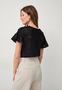 Schwarzes Spitzenshirt mit kurzen, rüschengesäumten Ärmeln, das ein kreisförmiges Ausschnittmuster aufweist. Getragen mit hoch taillierten beigen Hosen.