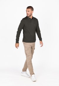 Polo a maniche lunghe verde scuro con colletto e tre bottoni, abbinato a pantaloni cargo beige e sneakers bianche.