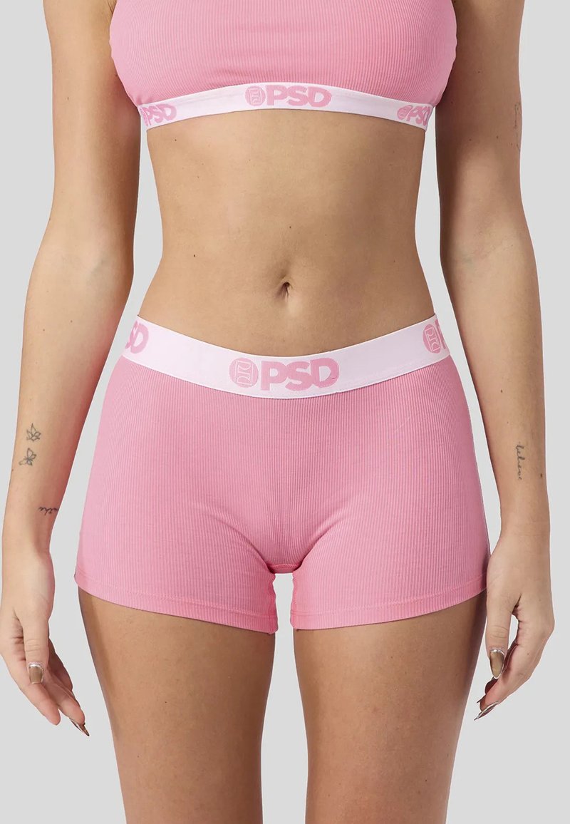 Femme portant un soutien-gorge de sport rose côtelé et un short assorti avec des bandes blanches à la taille marquées "PSD", debout avec les bras relâchés le long du corps.