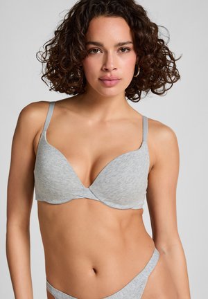 Vrouw met krullend haar, in een lichtgrijze bh en bijpassende onderbroek, recht vooruit kijkend tegen een effen grijze achtergrond.