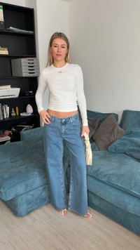 Jonge vrouw staand in de woonkamer, gekleed in een wit crop top met lange mouwen, wijde blauwe spijkerbroek, witte sandalen, en een creme scrunchie tas vasthoudend.