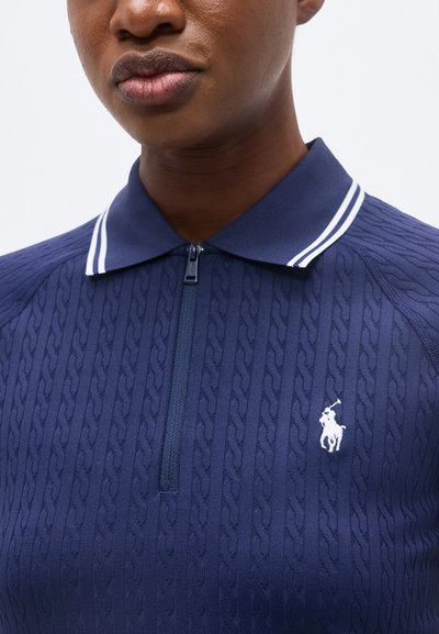 Κοντινή λήψη ενός ατόμου που φοράει μπλε ναυτικό πόλο με φερμουάρ και υφή, με λευκές ρίγες στο γιακά και λευκό λογότυπο αλόγου στο στήθος.