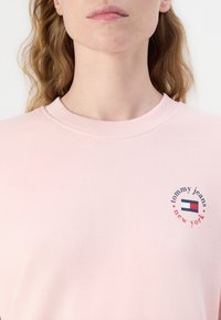 Felpa rosa chiaro con colletto a coste, caratterizzata da un logo circolare con il testo "tommy jeans" e un design di bandiera stilizzata sul petto.