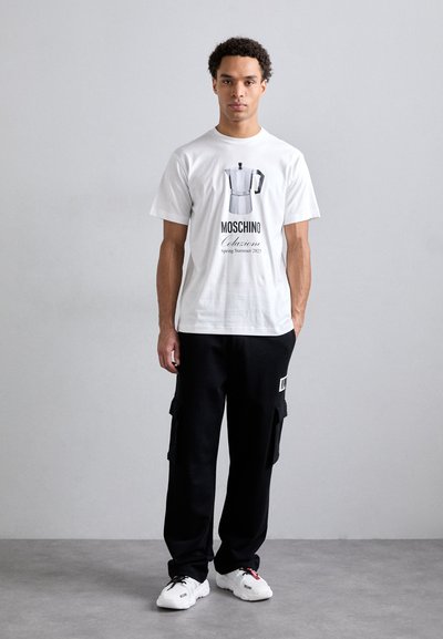 T-shirt blanc à manches courtes avec un graphisme de cafetière argentée, associé à un pantalon cargo noir et des baskets blanches. Design simple et moderne.