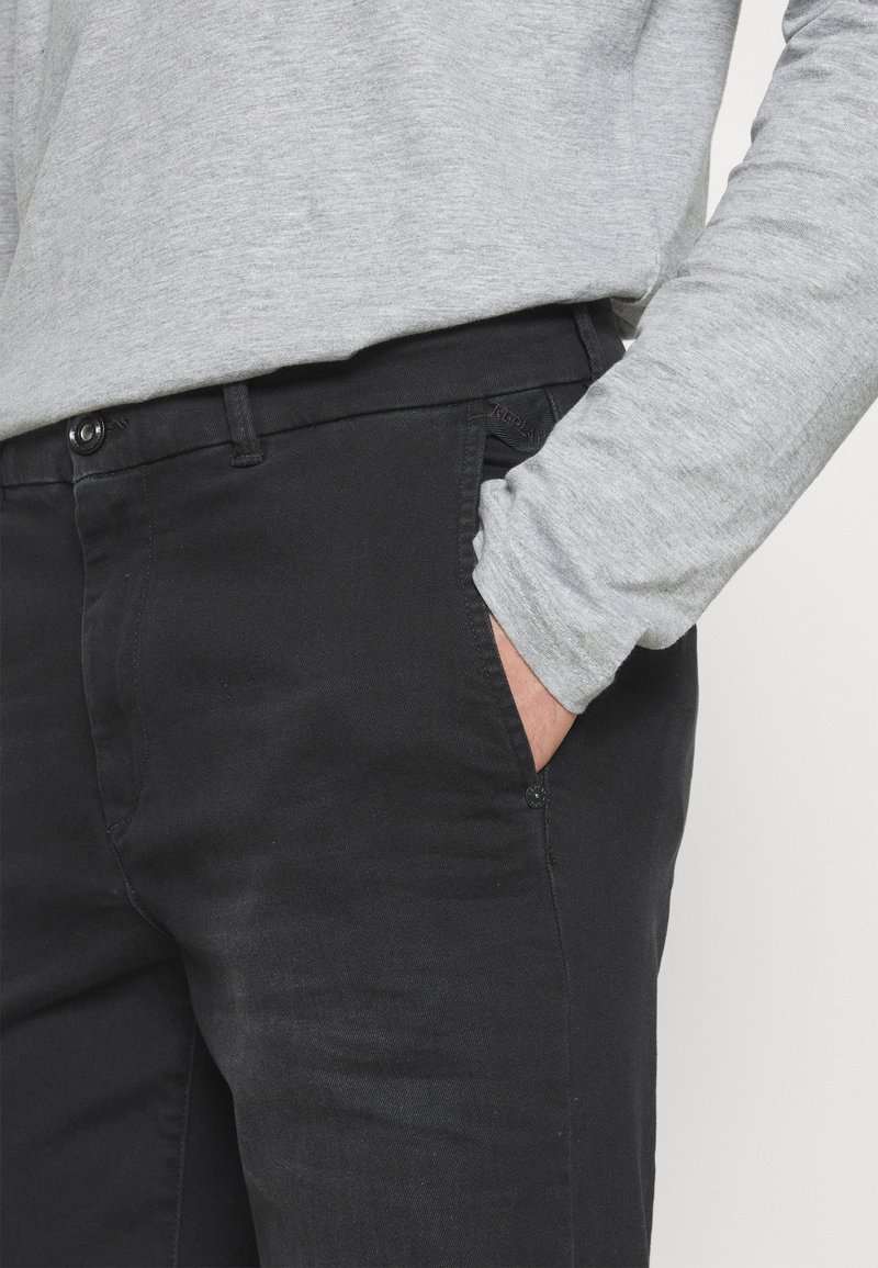 Schwarze Denim-Hosen mit seitlichen Taschen und Knopfverschluss, kombiniert mit einem hellgrauen Langarmshirt. Glatte Textur, figurbetontes Design.