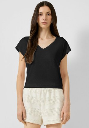 Frau mit langen braunen Haaren, die ein schwarzes V-Ausschnitt-Kurzarmtop und weiße strukturierte Shorts trägt, steht vor einem schlichten Hintergrund.