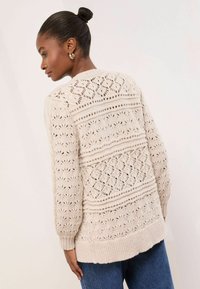 Beige kötött kardigán, texturált mintával, nyitott részletekkel, bordázott mandzsettával és egyenes szegéllyel. Lezser farmernadrággal kombinálva.