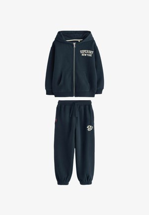 Temno modra majica s kapuco, z zadrgo spredaj in sprednjimi žepi. Ujemajoče se temno modre trenirke z elastičnim pasom in izvezenim logotipom.