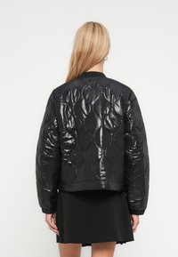 Γυναίκα με μακριά ξανθά μαλλιά, ντυμένη με λαμπερό μαύρο καπιτονέ μπουφάν και μαύρη φούστα, γυρισμένη με την πλάτη σε απλό φόντο.